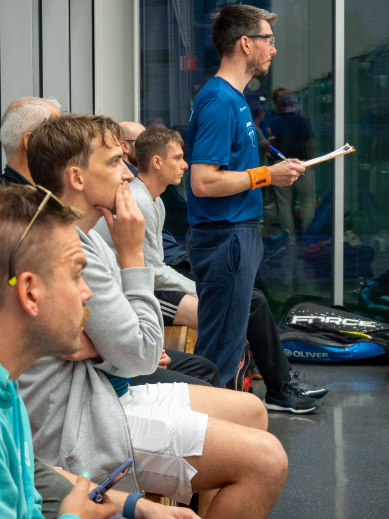 Spectators watching beside a squash court while a scorekeeper in a blue SCHIET shirt holds a clipboard.