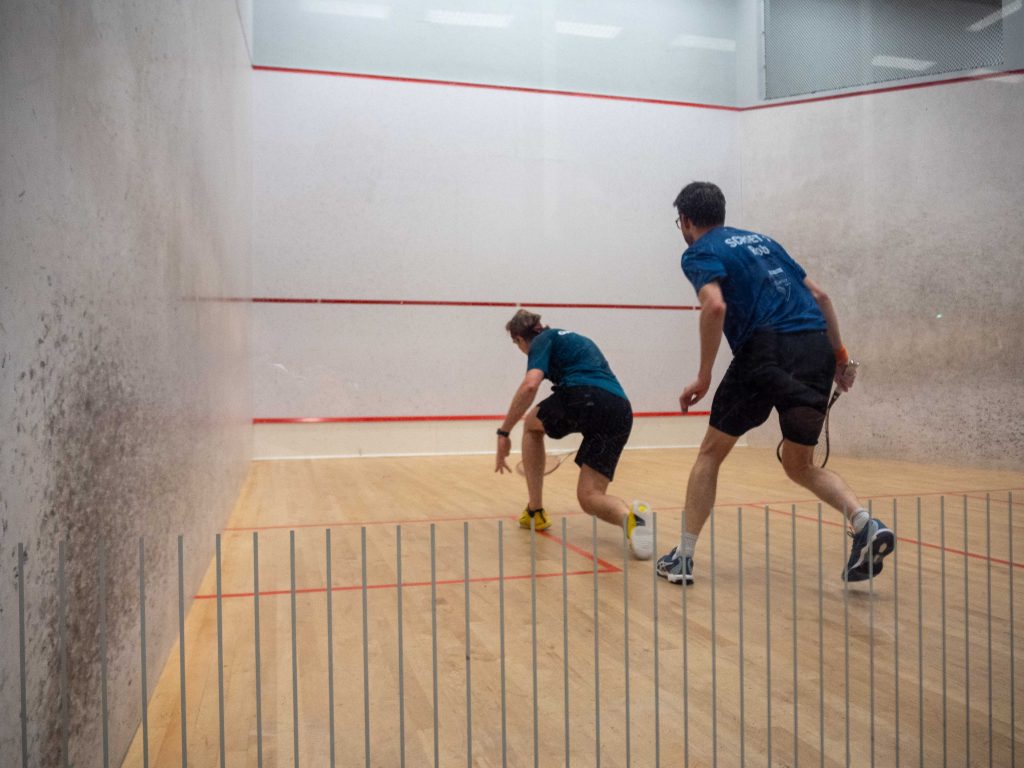 Two squash players contest a rally behind the glass court wall.
