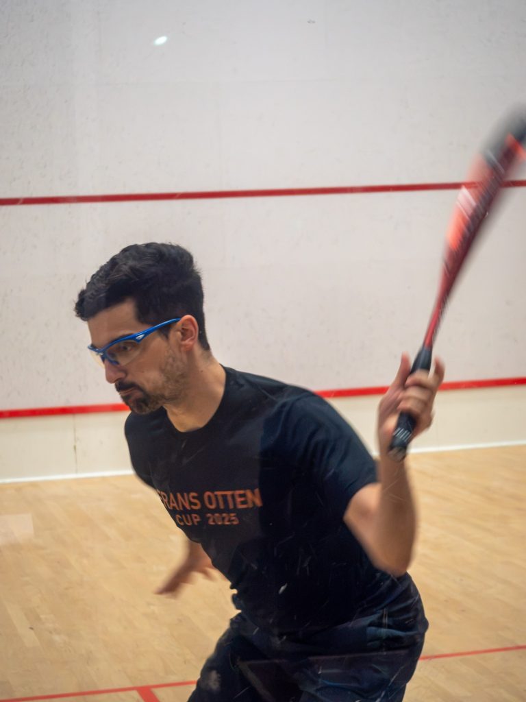 A squash player wearing blue protective glasses prepares a racket swing on court.