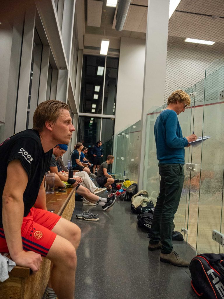 Spectators watching squash through the glass while a club member marks the score.