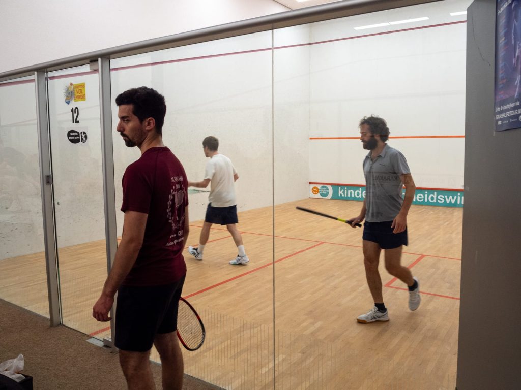 Players and a spectator beside the glass squash court during league week two.