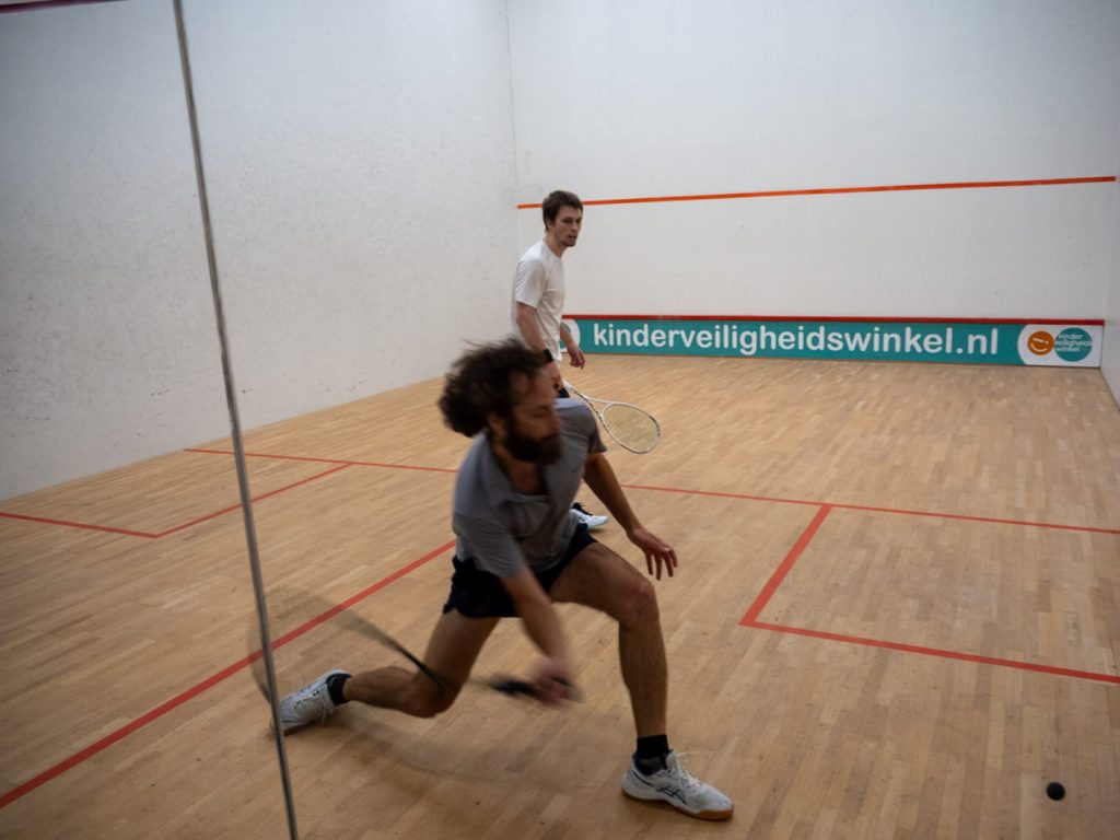 A player in a grey shirt lunges to reach the ball near the back wall.