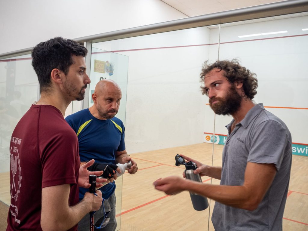 Three players talk beside the glass court between matches.