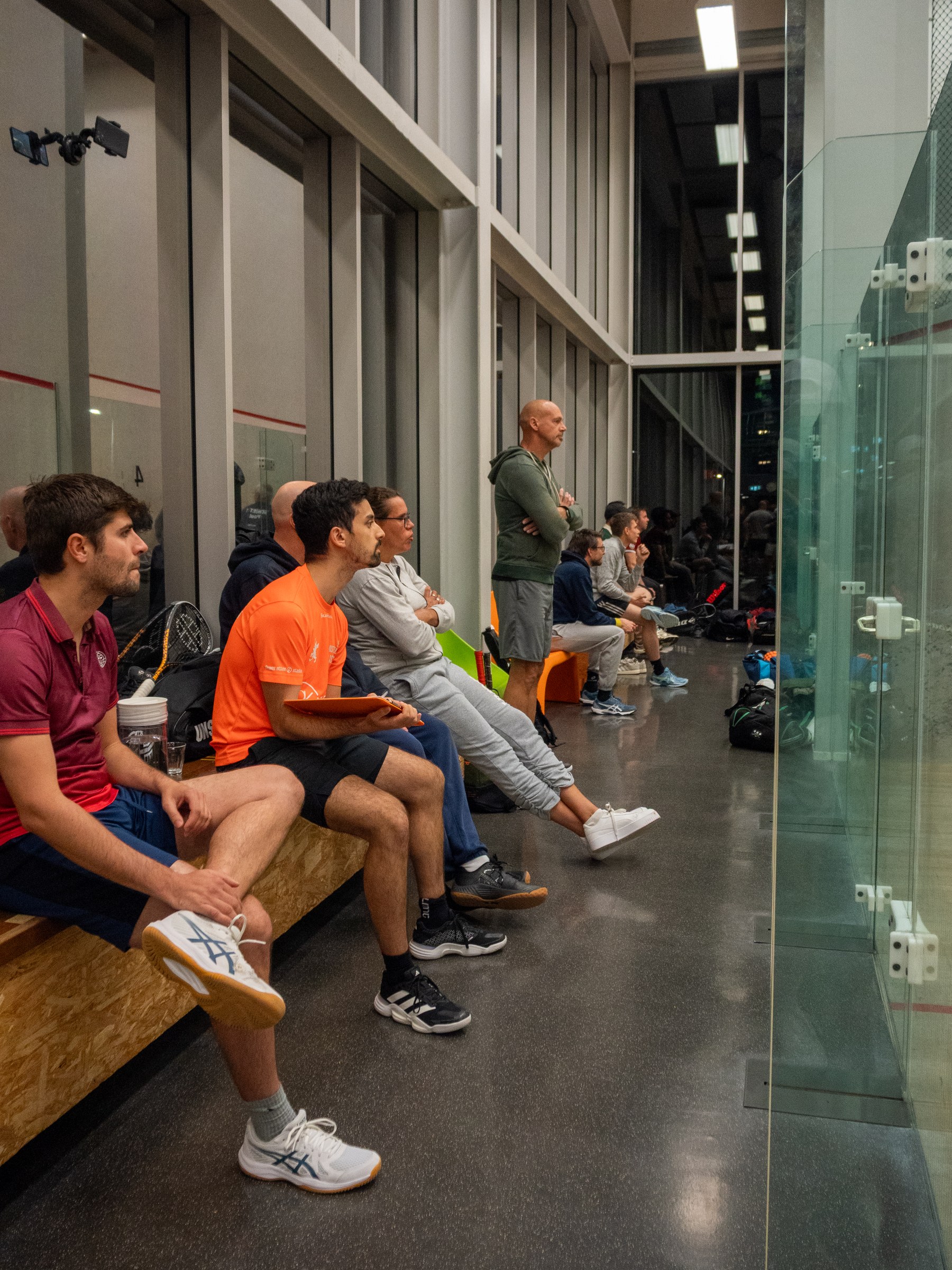 Players and spectators watching from the benches beside the glass courts.
