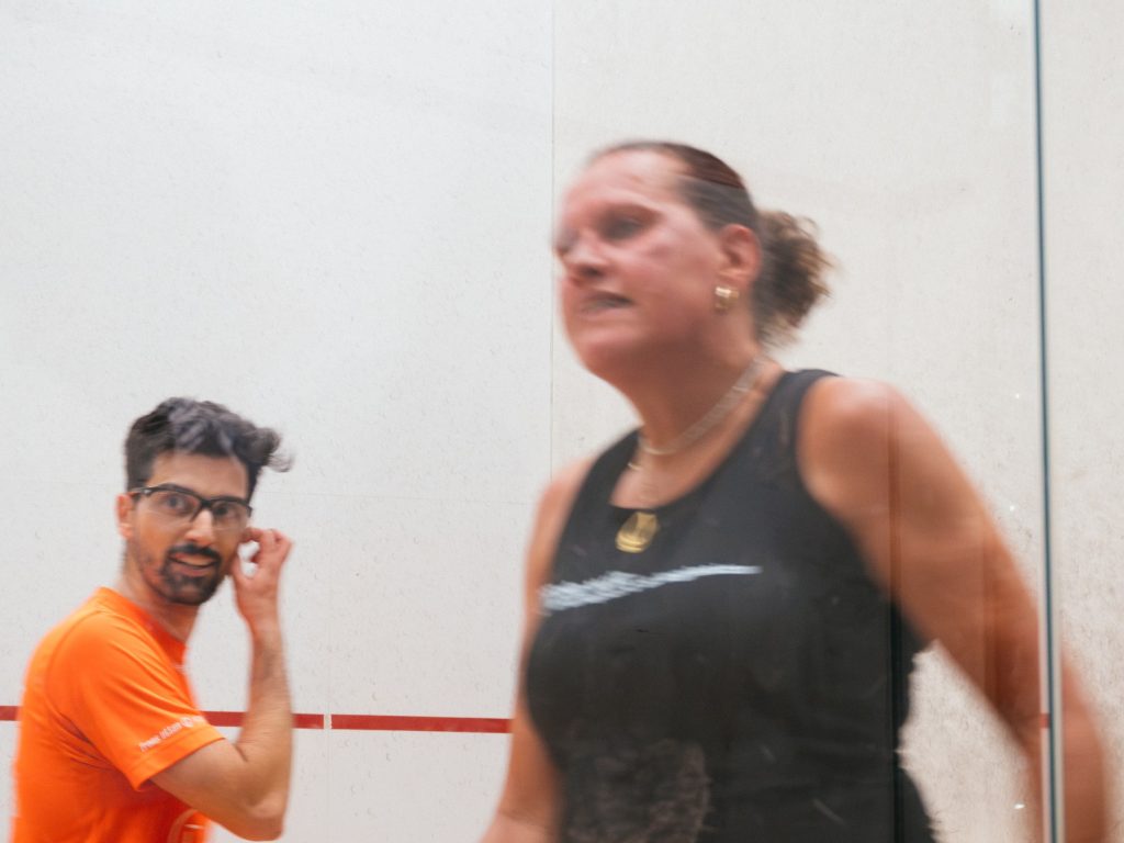 A player in black beside the glass court, with another player behind.