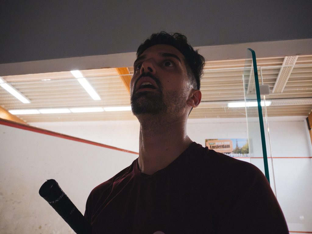 A player in a burgundy shirt holding a racket beside the glass squash court.