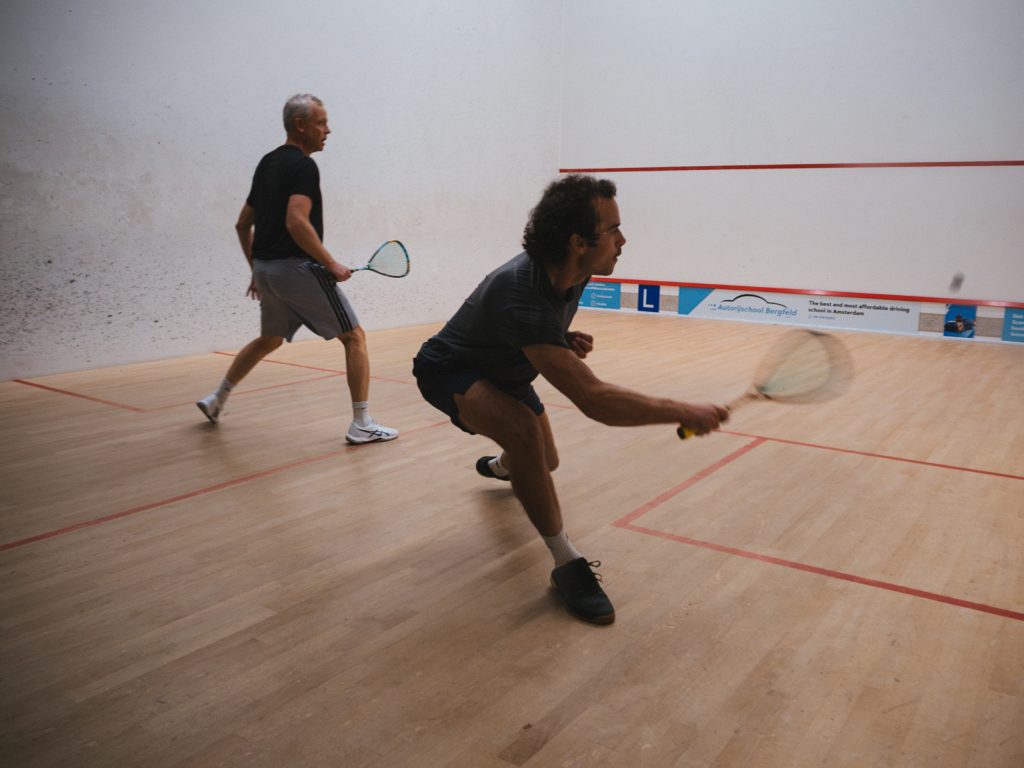 A player lunging forward to play the ball, with his opponent behind him on the squash court.